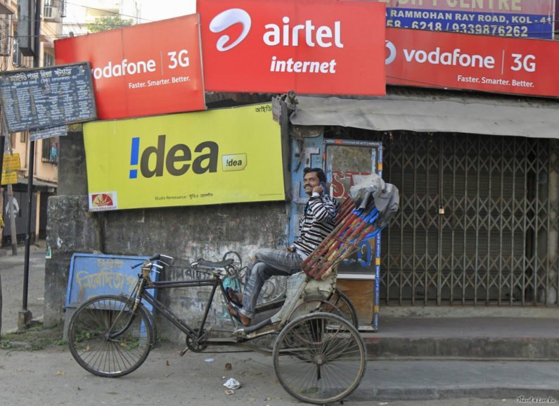 Велорикша с телефоном на фоне рекламы мобильных операторов Airtel, Vodafone и Idea