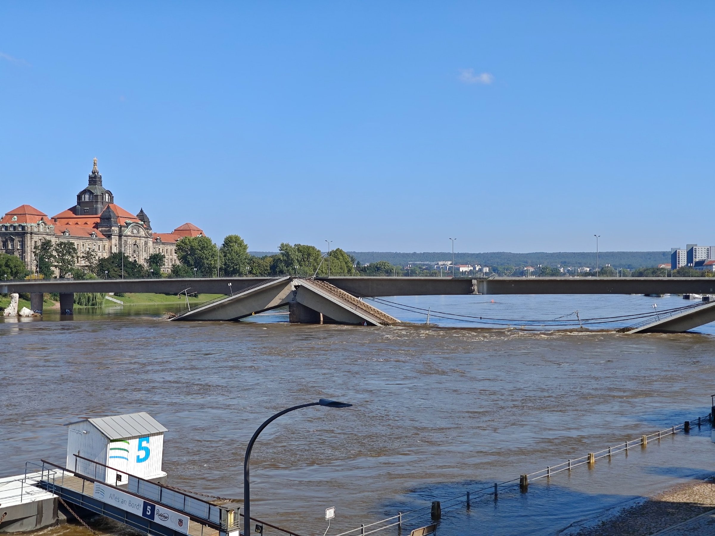 Мутная широкая река вровень с берегами, через неё мост, у которого средняя часть обрушилась в воду; на том берегу здание с куполом