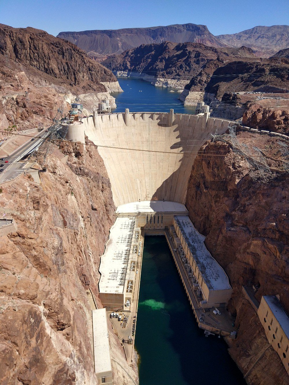 Изогнутая бетонная стена плотины между скалами, за ней водохранилище, внизу река