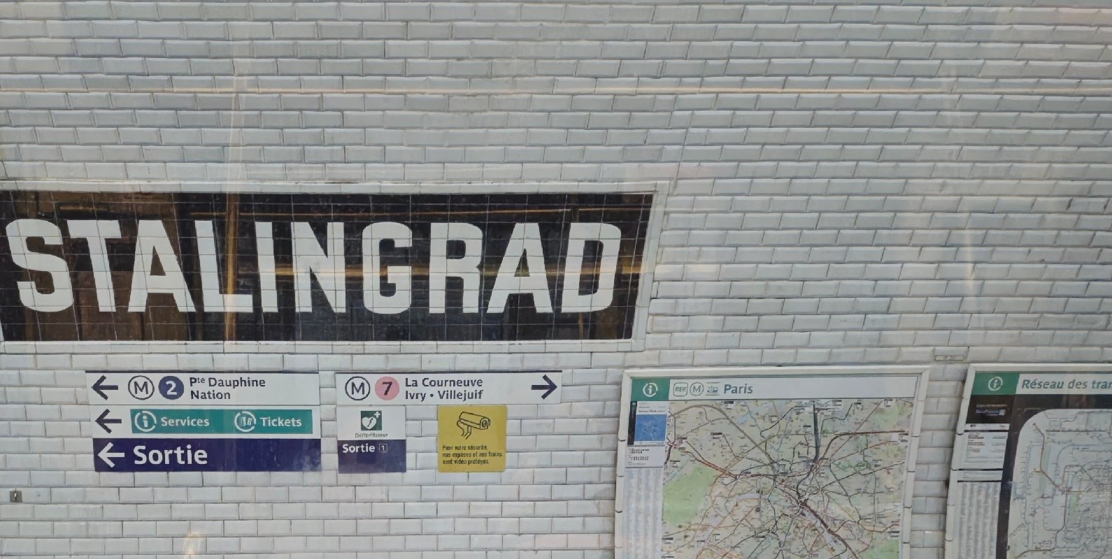 Белая кафельная стена станции метро: чёрная табличка STALINGRAD, указатели линий и схема метро