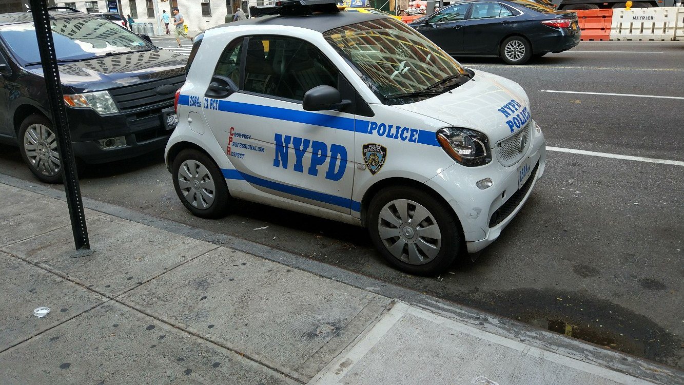 Крошечная бело-синяя полицейская машина с надписью NYPD у тротуара рядом с обычным внедорожником