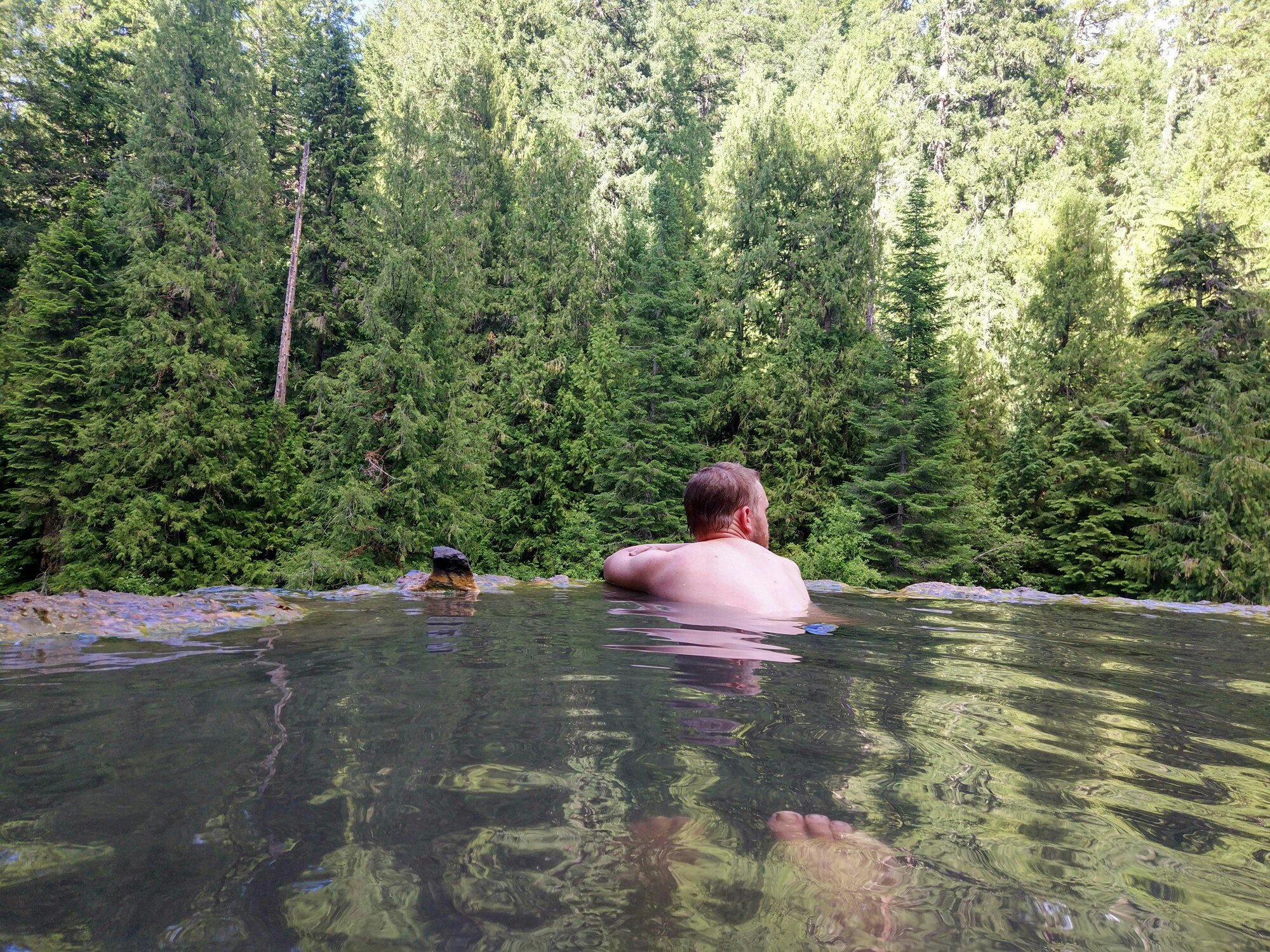 Павел сидит по плечи в прозрачной тёплой воде каменной чаши спиной к камере и смотрит на хвойный лес