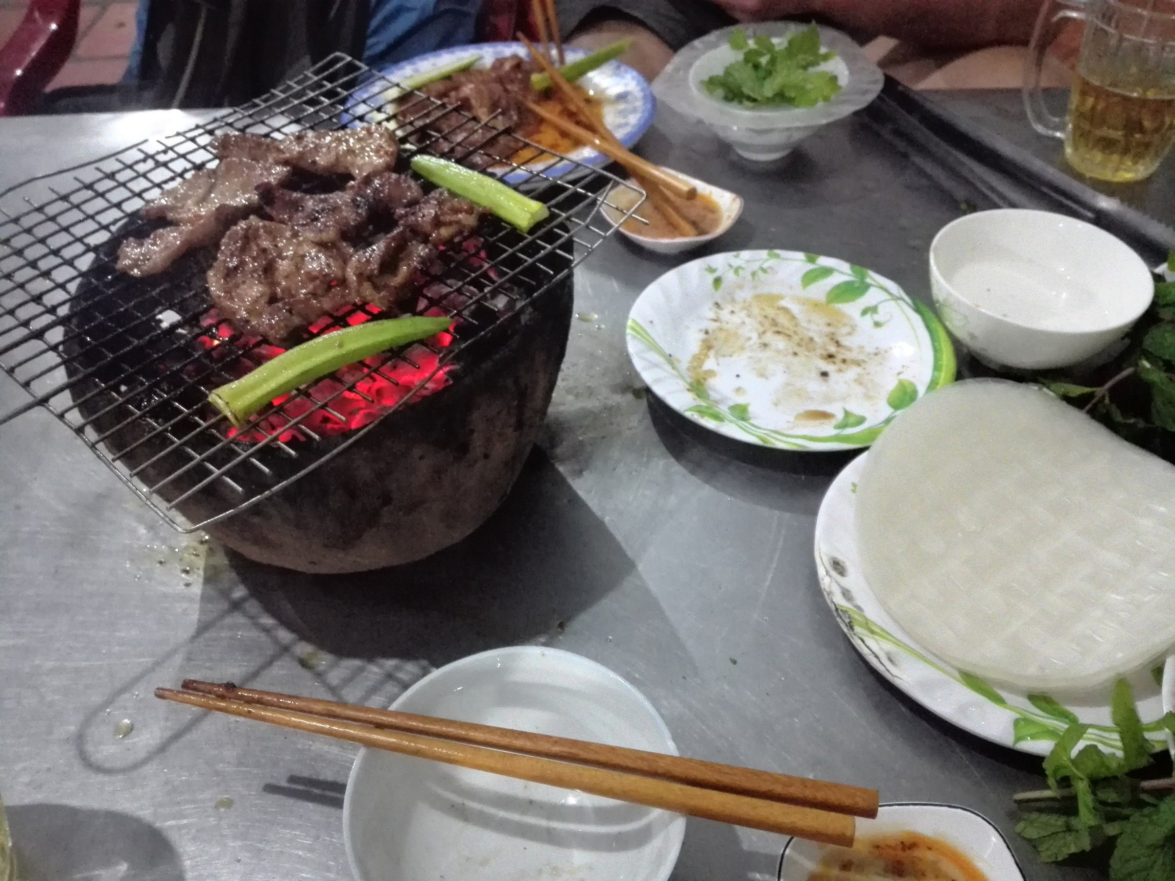 Мясо на решётке над углями в глиняной жаровне, рядом стопка рисовых листов и зелень