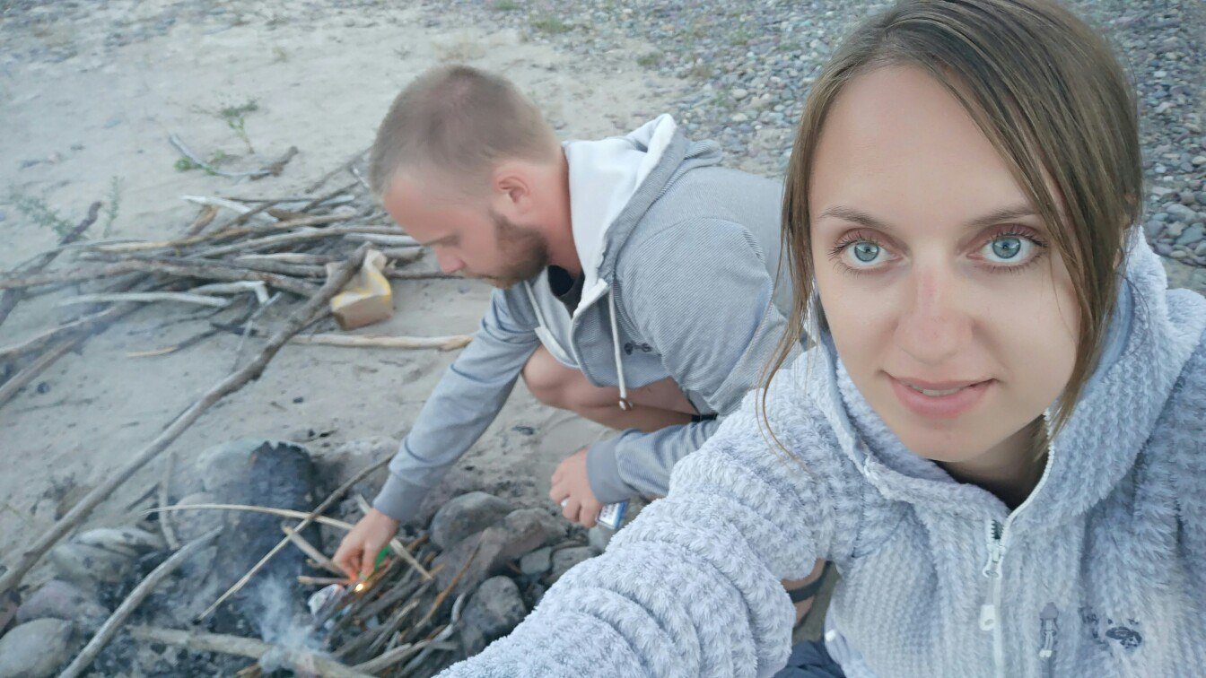 Павел разжигает костёр из сухих веток на галечном берегу, рядом Александра в светлой кофте делает селфи