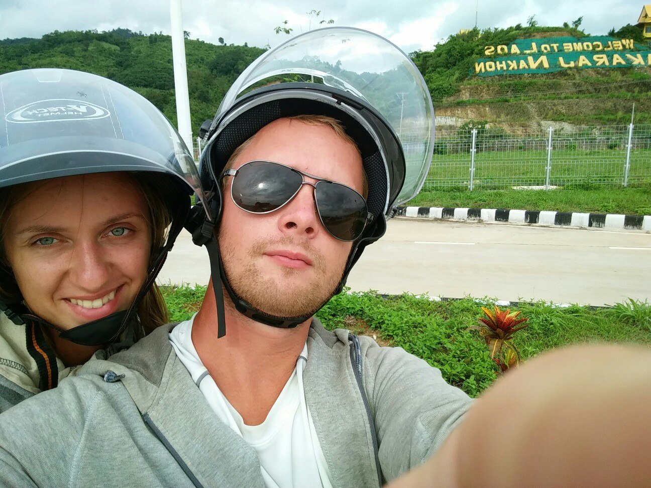 Павел и Александра в шлемах делают селфи у надписи Welcome to Laos на склоне