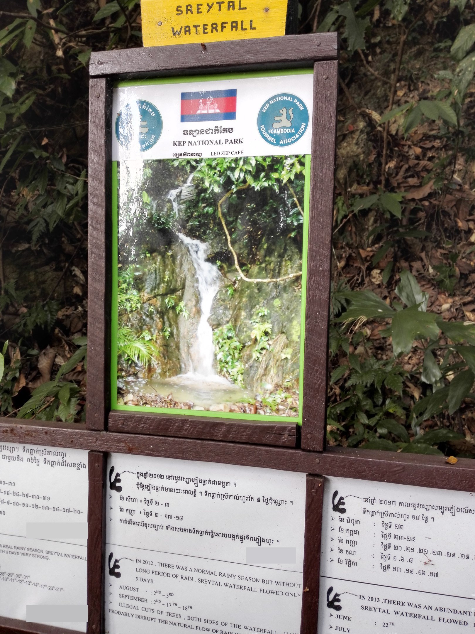 Стенд в лесу с фотографией водопада на скале и табличками на кхмерском и английском